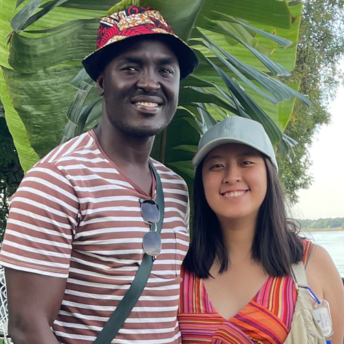 Kemish and his wife smile at the camera while standing in front of a large tropical plant with large leaves. 