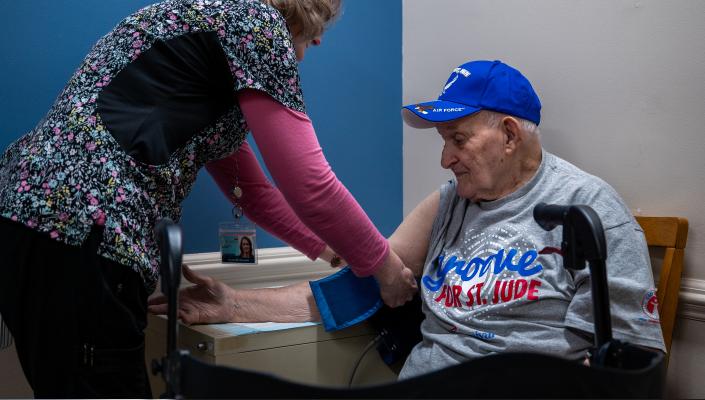 Frank Squibb, one of about 978 people participating in the ARIC study, gets his blood pressure taken by a nurse in colorful scrubs. Frank wears a blue baseball cap and a grey t-shirt.