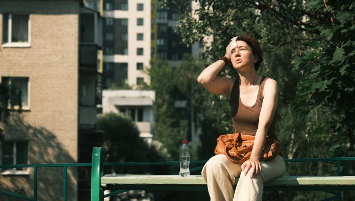 A woman sits on a city park bench while wiping her brow in the midday heat.