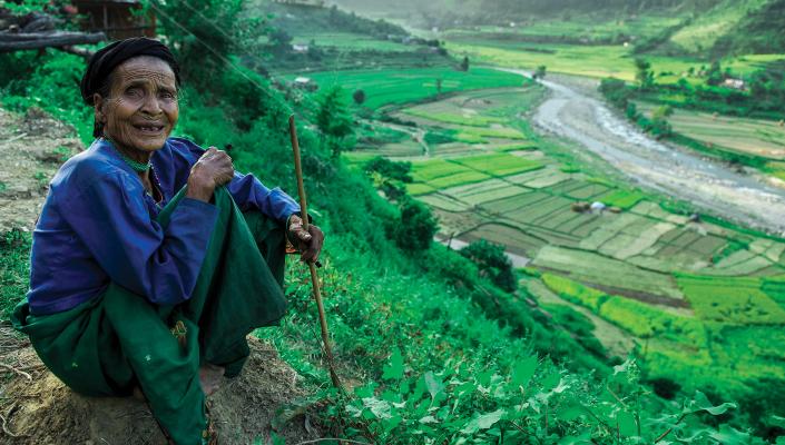 An older Nepali woman smiles and sits on a hill overlooking a field