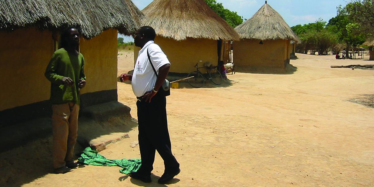 a CREATE worker talks with a community member outside a mud and thatch house