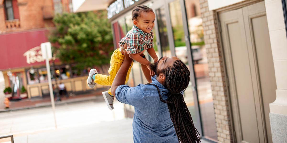 On a Baltimore street, a father lifts his young son up in the air. Both are smiling.