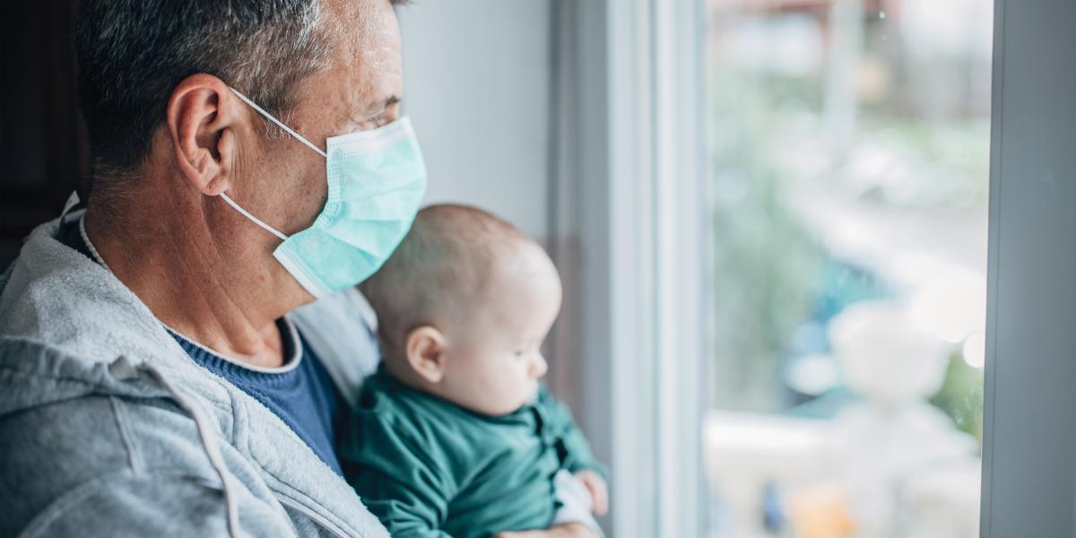 A man wearing a mask holds a baby while looking out of a window.