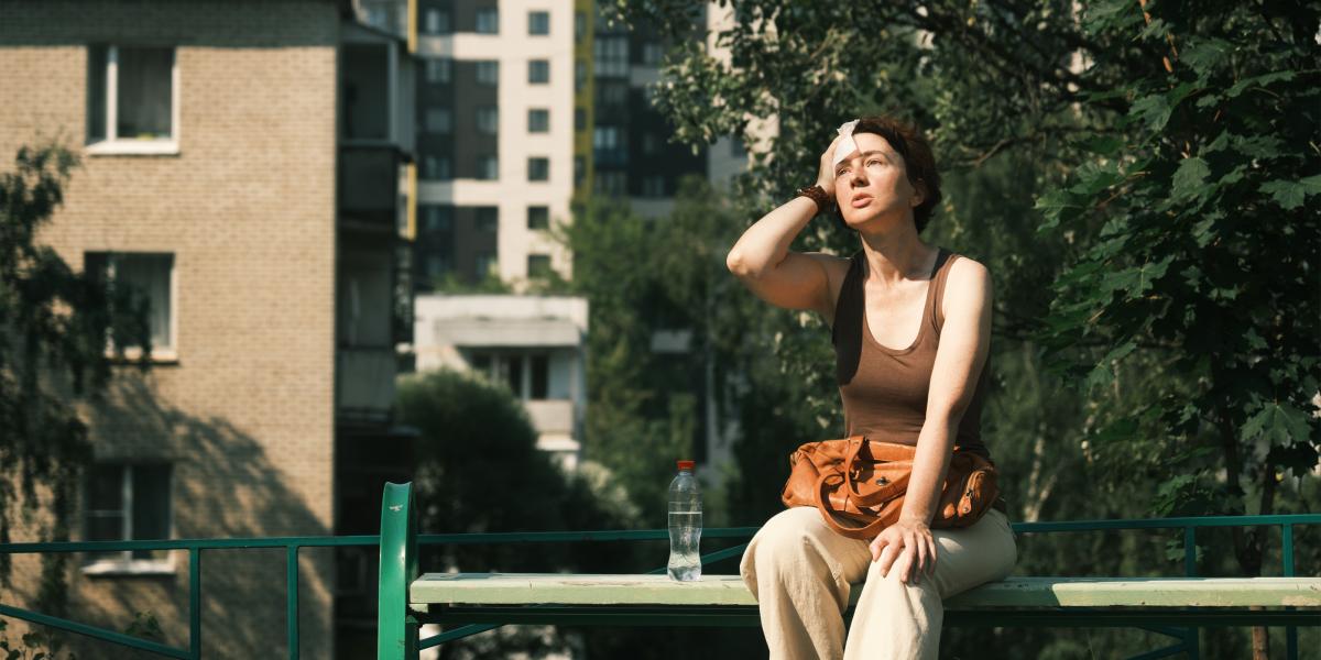 A woman sits on a city park bench while wiping her brow in the midday heat.