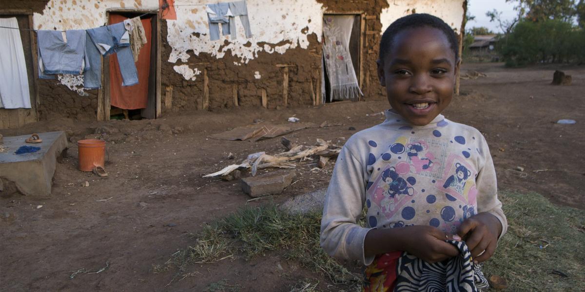 A child wearing a spotted shirt with teddy bears on itin her yard in Tanzania; behind her is her house and a clothesline with clothing hanging on it.