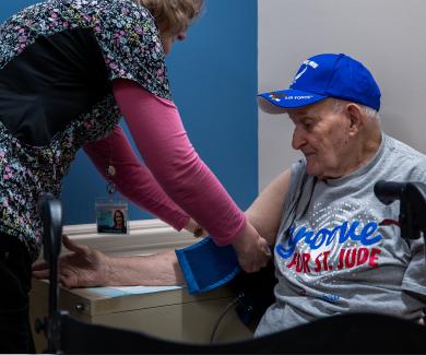 Frank Squibb, one of about 978 people participating in the ARIC study, gets his blood pressure taken by a nurse in colorful scrubs. Frank wears a blue baseball cap and a grey t-shirt.