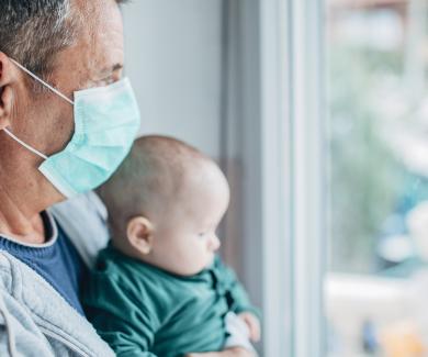 A man wearing a mask holds a baby while looking out of a window.