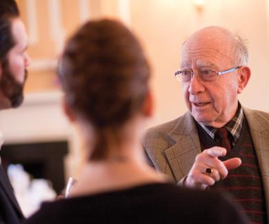 Clive Shiff, wearing a shirt, tie, and jacket speaks with a man and woman whose backs are to the camera