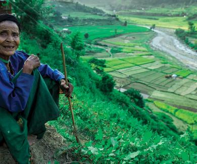 An older Nepali woman smiles and sits on a hill overlooking a field