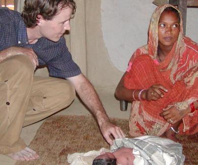 A researcher squats on the floor to examine a newborn infant; the child's mother, in a red sari, sits next to the infant.