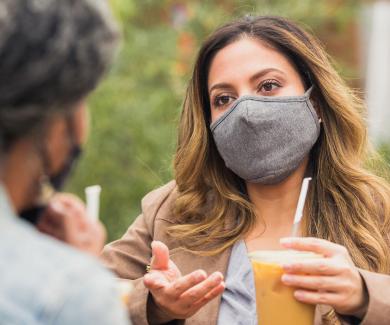 A woman wearing a mask speaks and gestures to a friend