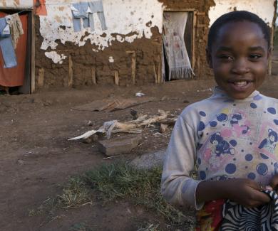 A child wearing a spotted shirt with teddy bears on itin her yard in Tanzania; behind her is her house and a clothesline with clothing hanging on it.